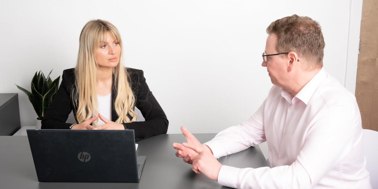 Office setting with Viktorija Thain in black blazer with laptop and colleague in white shirt at desk. Colleague is gesturing while Viktorija listens.