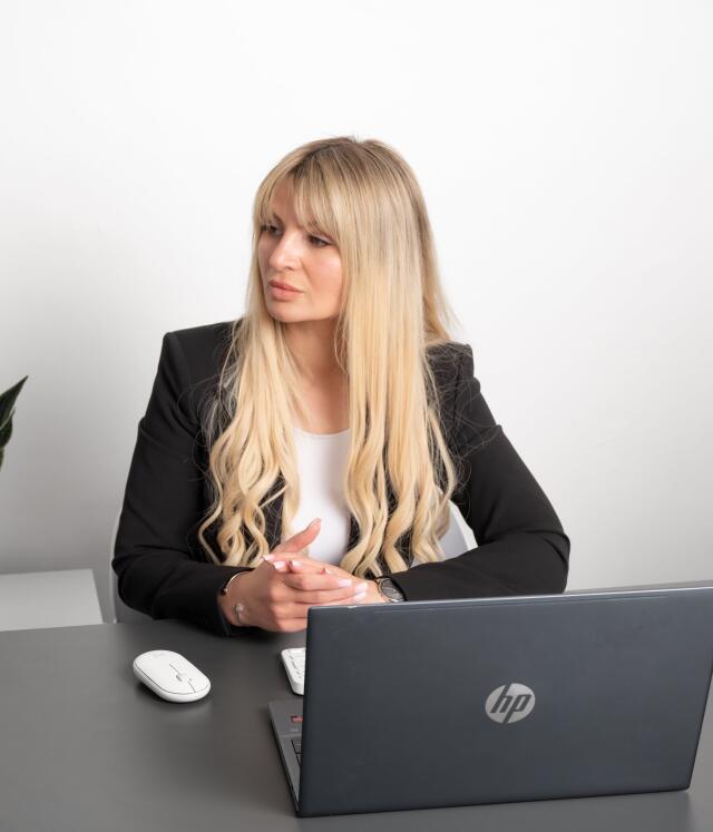 Viktorija Thain in black blazer sitting at desk with a laptop and mouse, looking to the side thoughtfully.