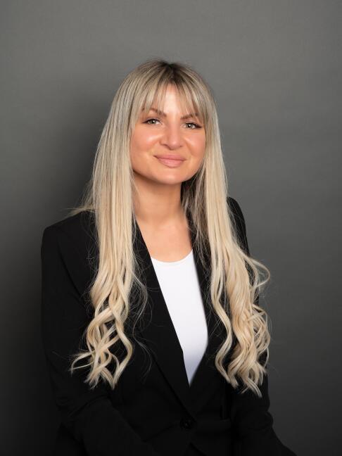 Portrait photo of Viktorija Thain smiling warmly, wearing black blazer over white top against gray background.