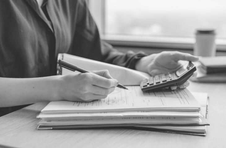 A person's hands hold a pen while writing in a notebook or binder on a wooden table. A calculator sits nearby to assist with any necessary calculations or budgeting.