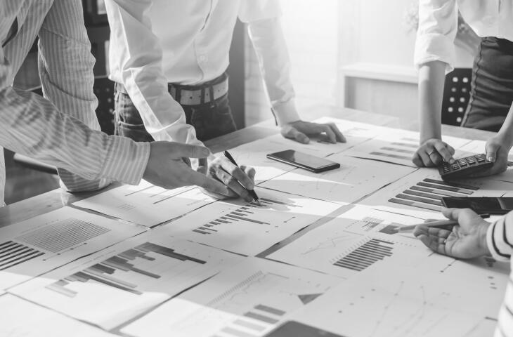 A group of businesspeople gather around a table covered in financial documents and charts. They point to and analyse various data points, using calculators and pens to make notes and calculations as they collaborate on a business plan or strategy.