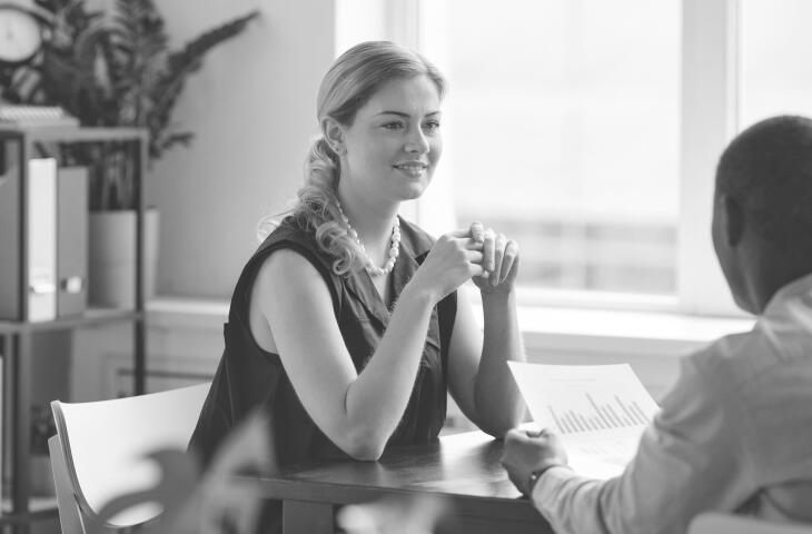 A young businesswoman wearing a black dress sits across a desk from a colleague, smiling as they engage in a friendly discussion. Plants and bookshelves decorate the warm, inviting office space, creating a pleasant atmosphere for their professional interaction.